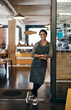 © HockleyM4/peopleimages.com - Happy woman, portrait or cafe with confidence for small business, entrance or welcome at indoor restaurant, Young, female person or waitress with smile, arms crossed or leaning on door at coffee shop