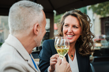 Smiling mature woman toasting with a white wine glass, enjoying a joyful moment with her husband at an outdoor restaurant, celebrating and sharing a special occasion with drinks and good company.