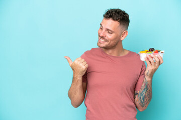 Wall Mural - Young caucasian man holding a bowl of fruit isolated on blue background pointing to the side to present a product