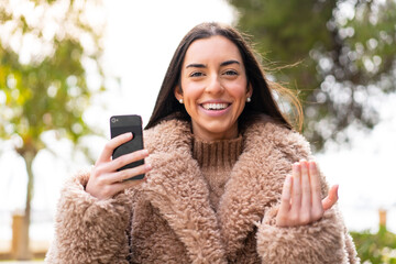 Young woman using mobile phone at outdoors inviting to come with hand. Happy that you came