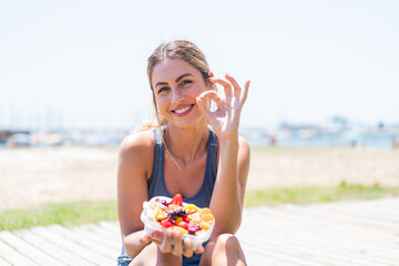 Wall Mural - Young pretty sport woman holding a bowl of fruit at outdoors showing ok sign with fingers