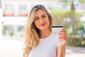 Canvas Print - Pretty blonde Uruguayan woman holding a credit card at outdoors smiling a lot