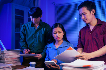 Three young businessmen work together in the office at night, busy on their laptops, checking documents, and finding solutions for an important meeting tomorrow.They are focused and working overtime