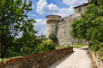 The pathway through Brescia castle park, highlighting the iconic Torre dei Prigionieri. Surrounded by green foliage, the historic tower and stone walls stand out under a picturesque sky