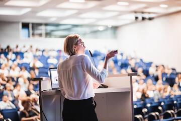 Female speaker giving a talk on corporate business conference. Unrecognizable people in audience at conference hall. Business and Entrepreneurship event