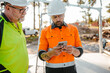 © Austockphoto - Two men wearing safety gear on the construction site with one holding a mobile device