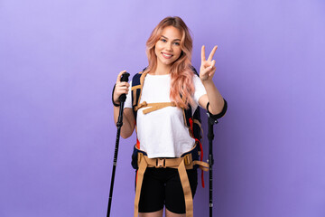 Wall Mural - Teenager girl with backpack and trekking poles over isolated purple background smiling and showing victory sign