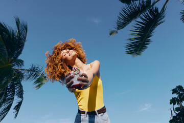 Woman joyfully holding a tropical drink against a blue sky backdrop with palm trees