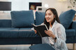 © amnaj - Young woman sits on the floor in a cozy living room, smiling happily while reading a book. The room is filled with natural light, creating a relaxing atmosphere for her leisure time