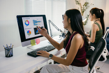 Two young businesswomen are analyzing charts and graphs on a computer screen, collaborating on a project in their bright and modern office