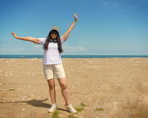 A beautiful young woman wearing a baseball cap and sunglasses on a sun-drenched beach expresses joy