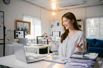 Young manager is having a video call on her laptop while working on marketing data in a modern office. She is smiling and gesturing while talking to her colleagues