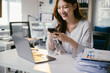 © PaeGAG - Young asian businesswoman is working in a modern office, using a smartphone and smiling, with a laptop and paperwork on her desk