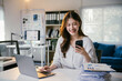 © PaeGAG - Young asian businesswoman multitasks with a smartphone and laptop in a modern office, focusing on data analysis, marketing, finance, and accounting
