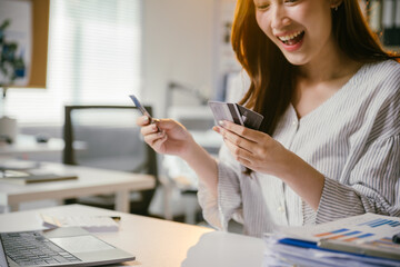 Cheerful young businesswoman happily managing finances with credit cards at her desk in a busy office