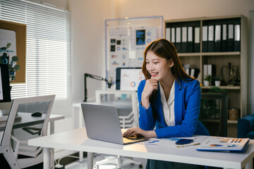 Young asian businesswoman smiling and working on laptop in bright office with blue window blinds. She looks happy, focused, and confident surrounded by charts and paperwork