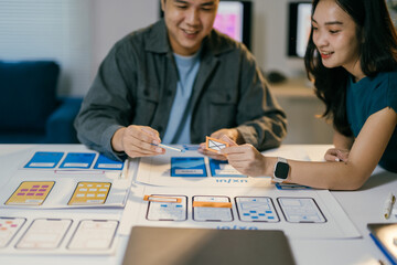 Two asian ui/ux designers are working together in office, holding ui element icons and discussing about mobile app interface wireframe design on table