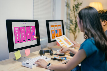 Two designers are working on a new mobile application in an office environment, comparing paper wireframes to a digital prototype displayed on a desktop computer