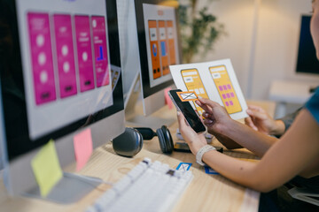 Two female ui/ux designers are discussing user interface design for a mobile application, holding a smartphone and paper prototypes in their hands