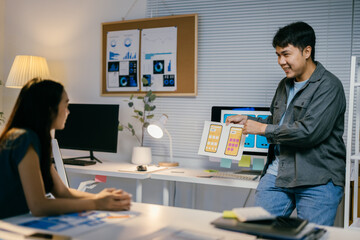 Two asian ui/ux designers collaborate late at night in an office. Discussing mobile app interface design