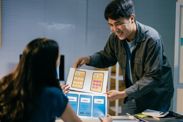 two asian ui/ux developers are discussing new mobile application wireframe designs, working late at 