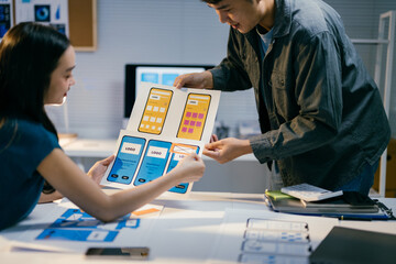 Two asian ui ux designers working together in office, carefully reviewing mobile app interface wireframe design on paper
