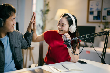 Two young content creators enthusiastically high-fiving while recording a podcast in their cozy home studio, surrounded by microphones and headphones, showcasing teamwork in content creation