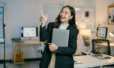 Young manager is holding a laptop and pointing with her finger while explaining a business strategy to her colleagues in a modern office