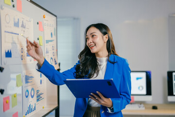Fototapete - Young asian businesswoman is holding a folder and pointing at charts on a whiteboard while smiling. She is giving a presentation to her colleagues in the office