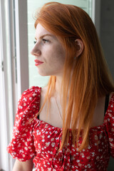 Red-haired young active girl with a smile on her face in a red dress. Classic portrait by the window.