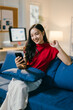 © Parichat - Young asian woman sits happily on a blue sofa in her modern home office, using her smartphone in the cozy, natural-lit room