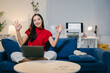 © Parichat - Young asian freelancer is sitting on a blue couch in her living room, working on her laptop and showing the ok sign with both hands