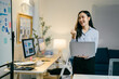 © Parichat - Young businesswoman is holding a laptop and smiling while standing in a modern office with graphs and charts displayed on the walls and computer screens