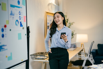 Young asian businesswoman is using her smartphone and smiling while standing in a modern office with a whiteboard in the background