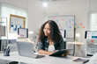 © PaeGAG - Young african american manager is sitting at her desk and working on a laptop while holding a report in her other hand. She is smiling and looking at the laptop screen
