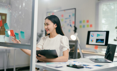 Young manager is smiling while planning a project using adhesive notes on a glass wall in her office, she is holding a clipboard and sitting at her desk