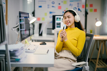 Businesswoman taking a break from work, sitting at office desk and listening to music. She is smiling and holding a pen in her hand