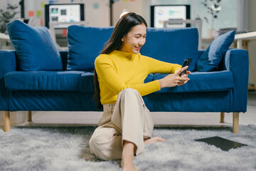 Asian woman taking a break in her modern home office, checking her phone and relaxing on a stylish sofa. She looks happy and content in this cozy setting, balancing work and leisure