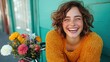 © JoxyAimages - A smiling woman wears a cozy sweater and sits by a bouquet of vibrant flowers, capturing a lively moment full of warmth and cheer against a green wall.