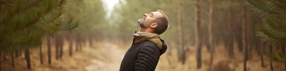 A man is standing in a forest, looking up at the sky. He is wearing a black jacket and a brown scarf. Scene is peaceful and serene, as the man is taking a moment to appreciate the beauty of nature