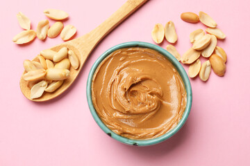 Wall Mural - Tasty peanut butter in bowl and spoon with groundnuts on pink table, flat lay