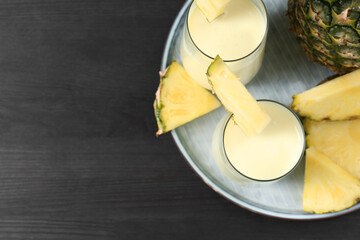 Tasty pineapple smoothie in glasses and fruit on black wooden table, top view. Space for text