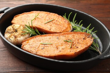 Halves of tasty cooked sweet potato with rosemary and garlic in baking dish on wooden table, closeup