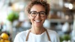© Pinklife - A cheerful woman with glasses and short curly hair wearing a white apron, smiling at the camera in a vibrant market setting filled with fresh produce.