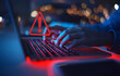 © Piya W. - A person types on a laptop, illuminated in red, with a warning symbol highlighted, indicating a potential cybersecurity issue.