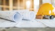 © Vilaysack - Rolled-up blueprints on a construction site table with a yellow hard hat, representing architecture and building planning.