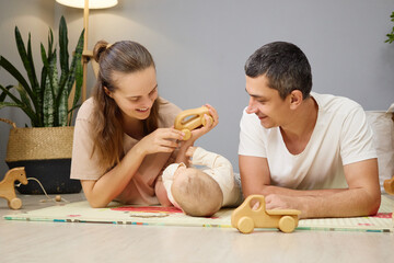 Smiling cheerful woman and man lying on floor with infant interacting with toddler child together enjoying babyhood moments of care and development in nursery room at home