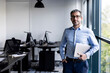 © Westend61 - Smiling businessman standing with laptop near window in office