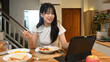 © Prathankarnpap - Smiling young woman sitting at a kitchen table, enjoying meal and watching something on digital tablet