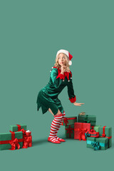 Beautiful young woman dressed as elf with Christmas gifts showing silence gesture on green background